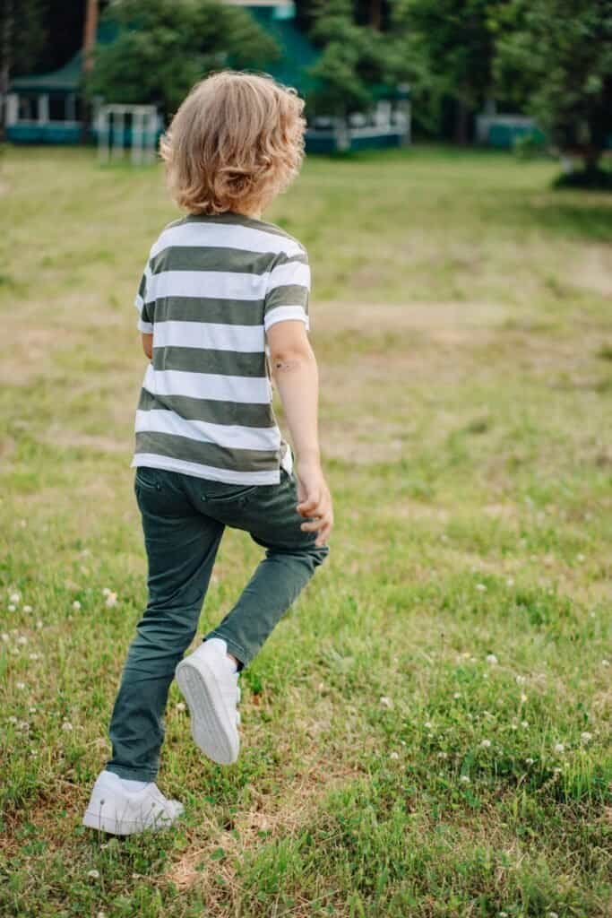 Young child walking outdoors in a park with green grass and trees.